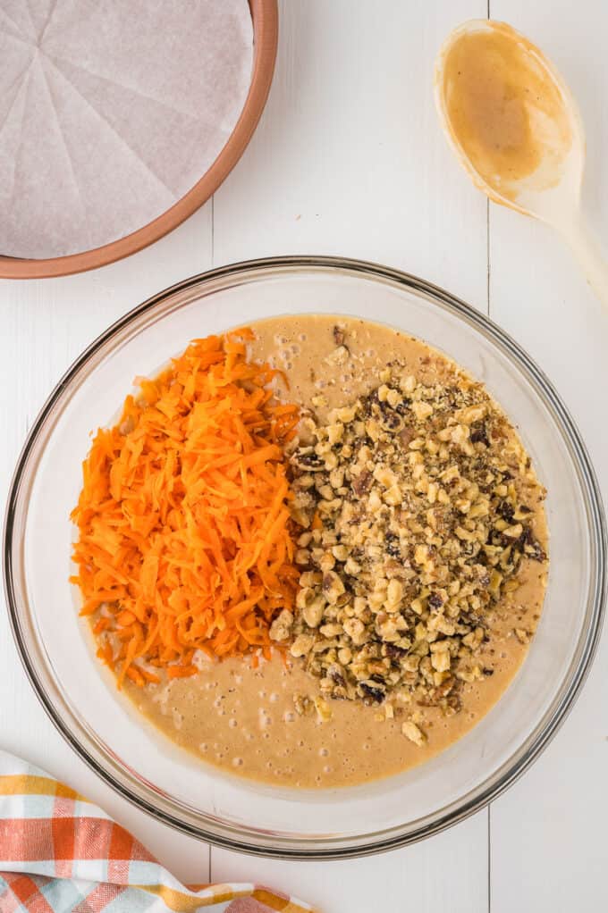 a glass bowl of cake batter with shredded carrots and walnuts.