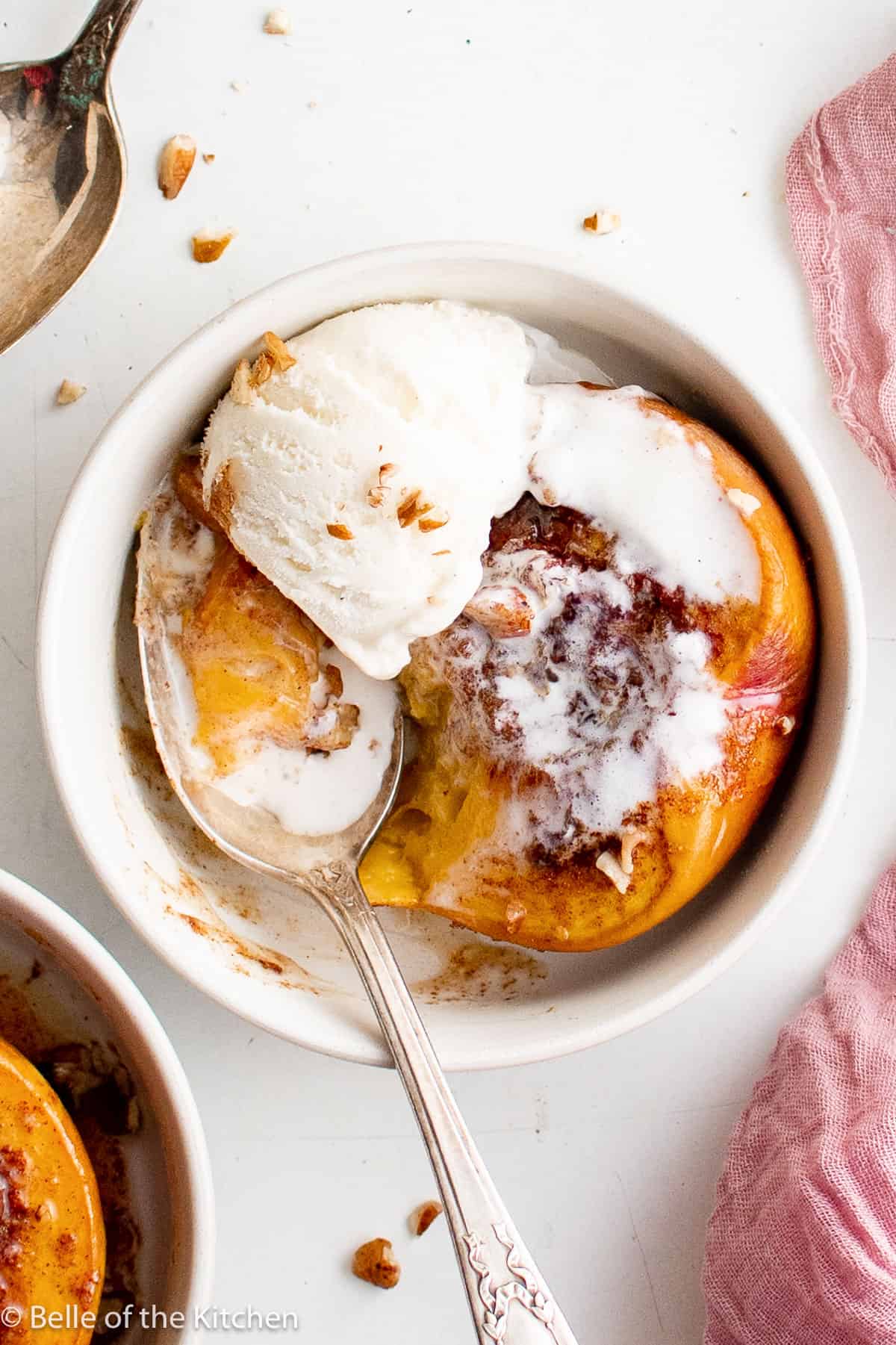 a roasted peach with ice cream in a white bowl being scooped out with a spoon.