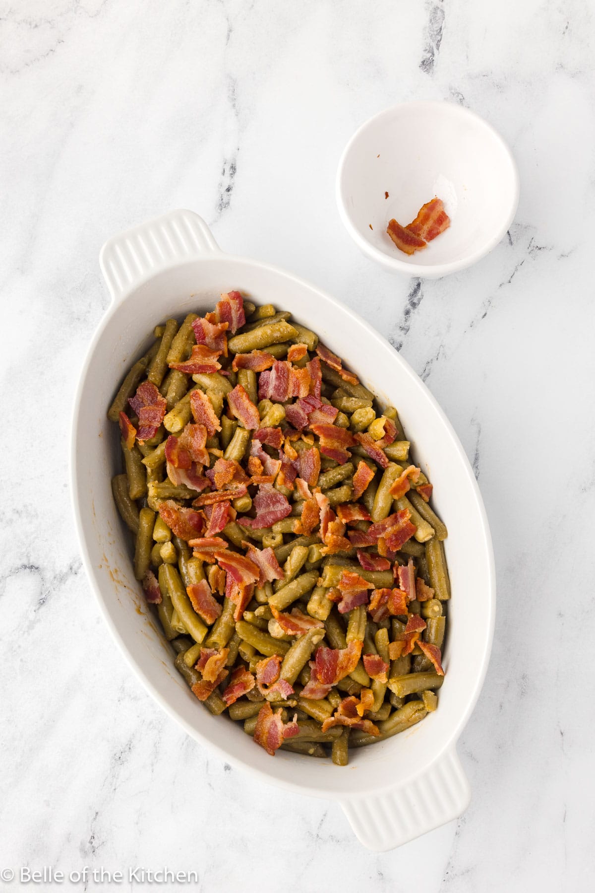 a white baking dish full of cooked green beans topped with bacon pieces.