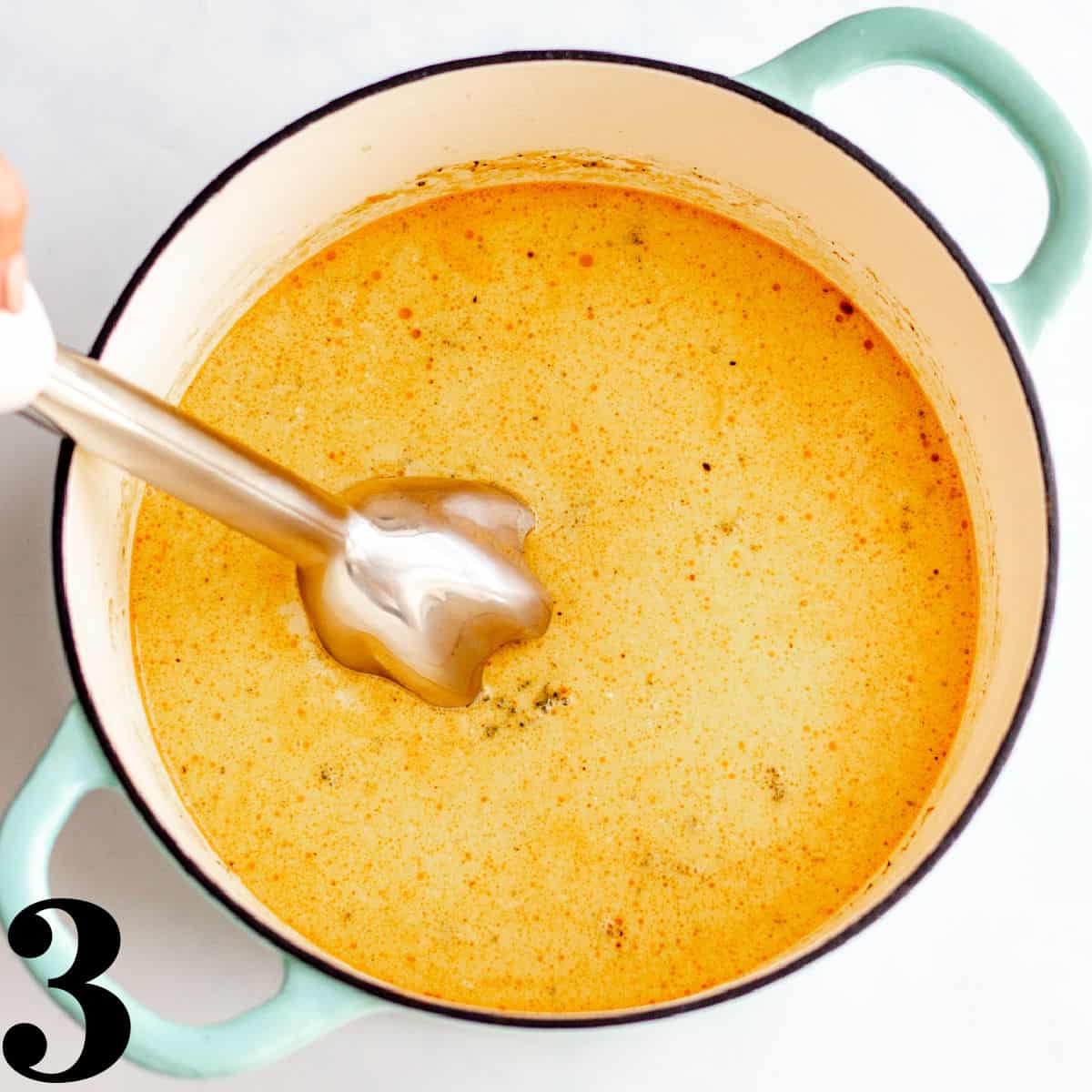 A dutch oven full of broccoli cheddar soup with an immersion blender.