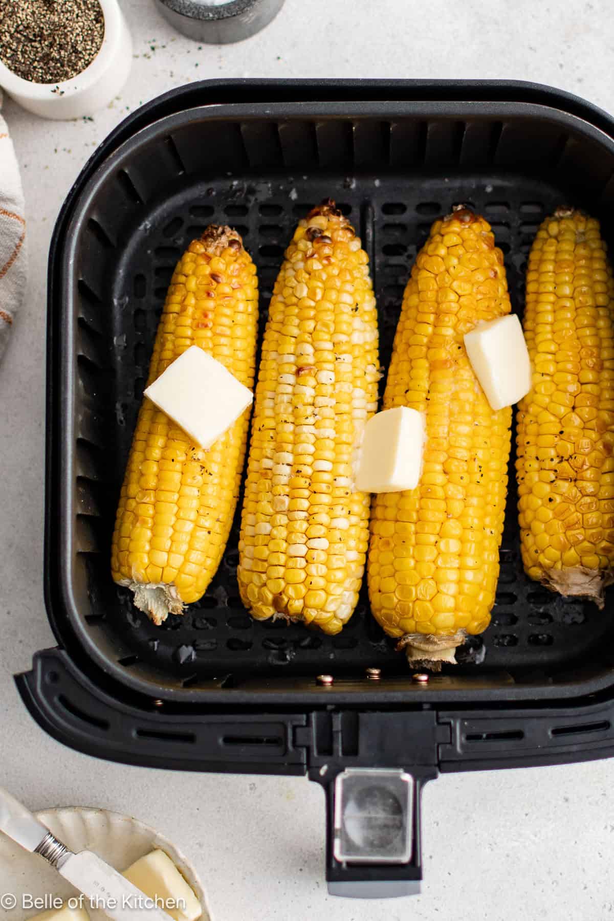 Ears of corn inside of an air fryer basket with pats of butter on top.