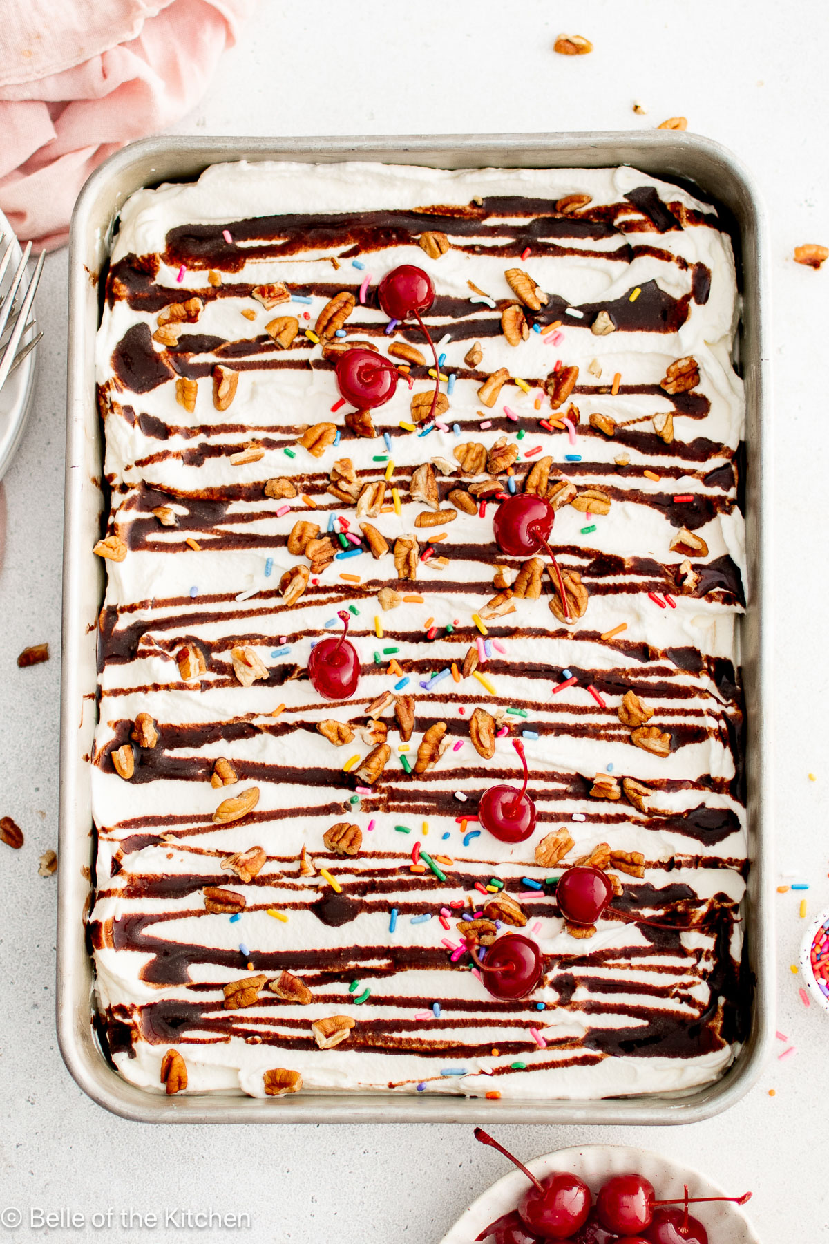 A cake pan full of banana split cake topped with chocolate drizzle, sprinkles, nuts, and cherries.