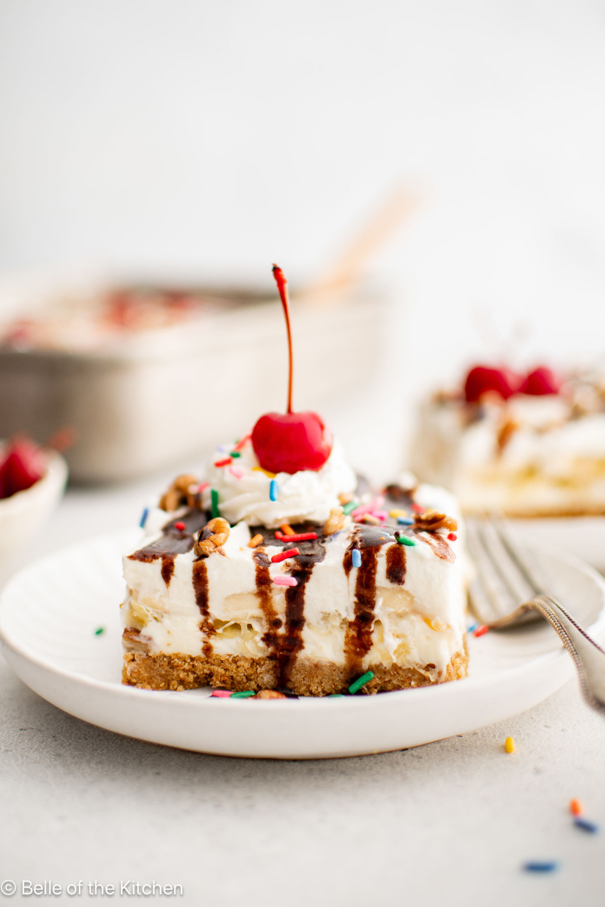 A slice of banana split cake on a plate with a cherry on top.