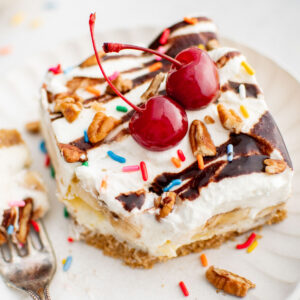 A slice of banana split cake on a plate with cherries on top next to a fork.