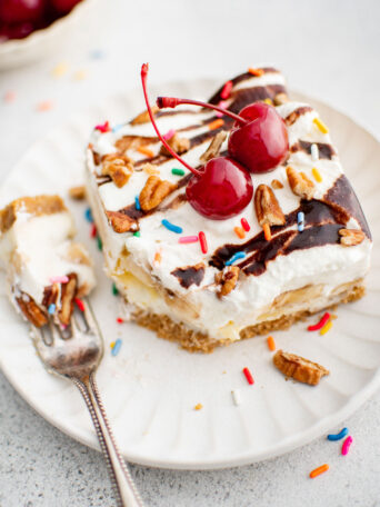 A slice of banana split cake on a plate with cherries on top next to a fork.