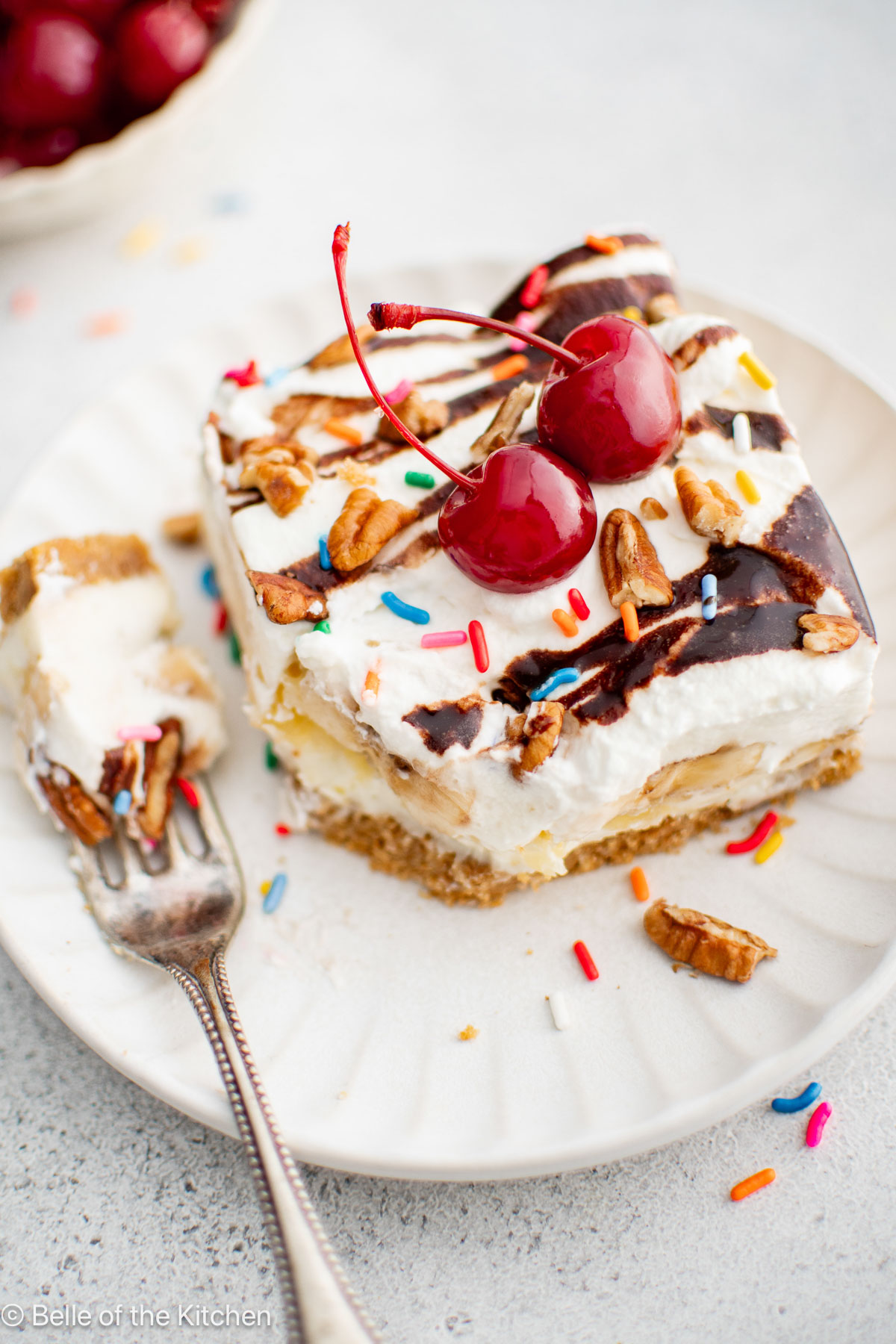 A slice of banana split cake on a plate with cherries on top next to a fork.