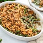 Green bean casserole in a white baking dish with fried green onions on top and a spoon scooping some out.