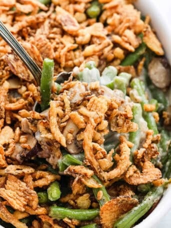 Close up shot of a spoon scooping up green bean casserole from a white baking dish.