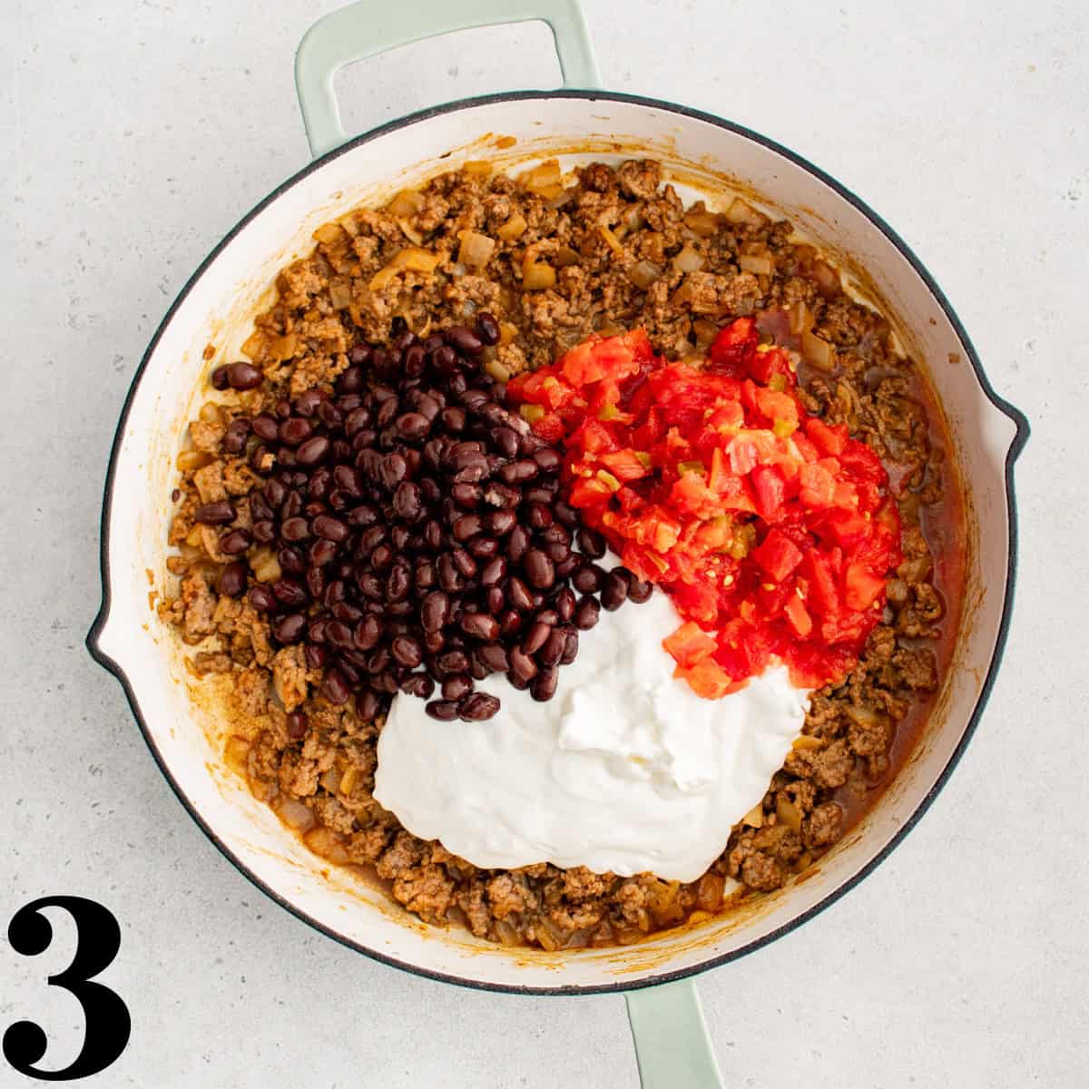 Cooked beef in a skillet with black beans, tomatoes, and sour cream on top.