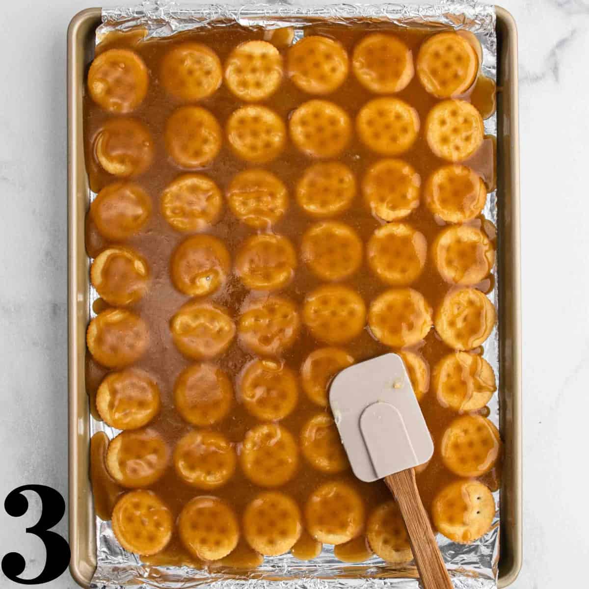 Round crackers laid out on a cookie sheet with caramel sauce on top with a spatula.