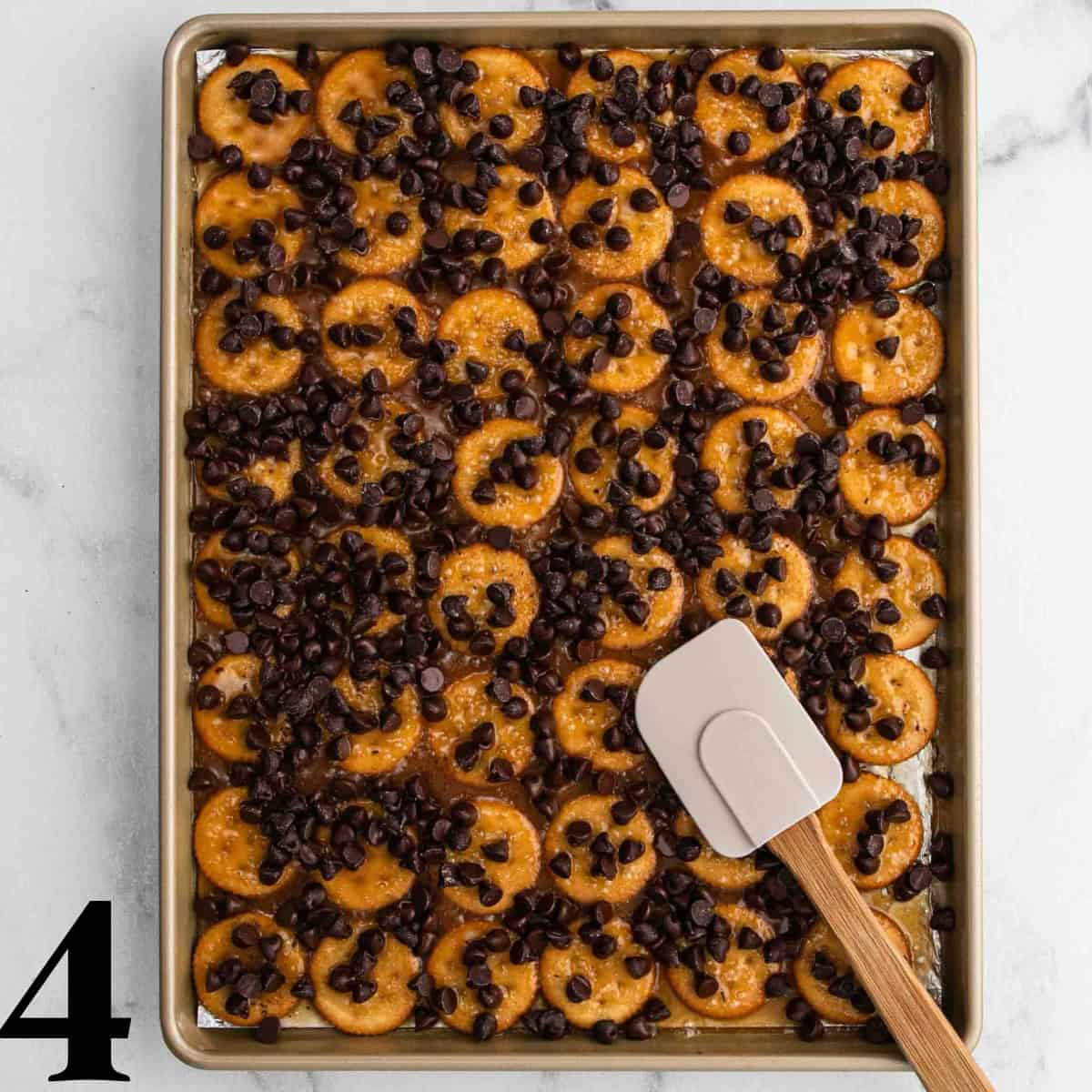 Round crackers laid out on a cookie sheet with chocolate chips on top with a spatula.