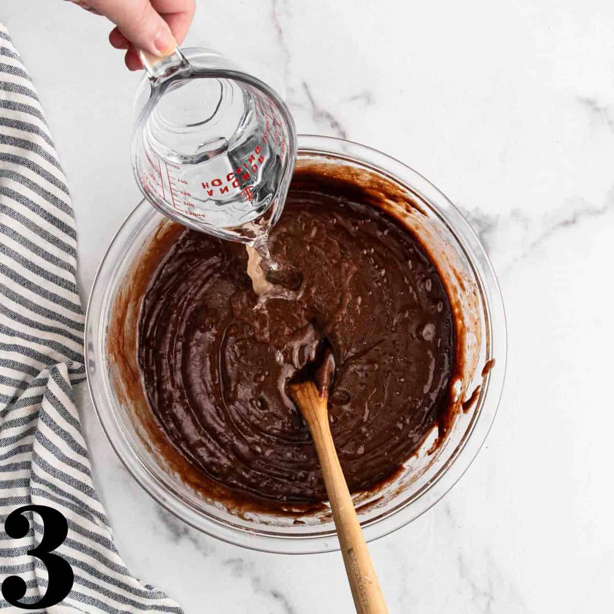 A glass bowl of chocolate cake batter with water being poured into it.