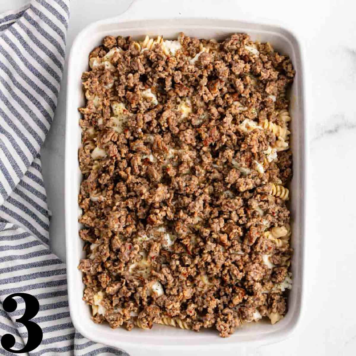 Ground sausage on top of pasta in a white baking dish.