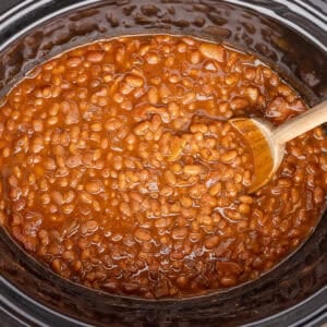 A black crockpot full of baked beans and a wooden spoon.