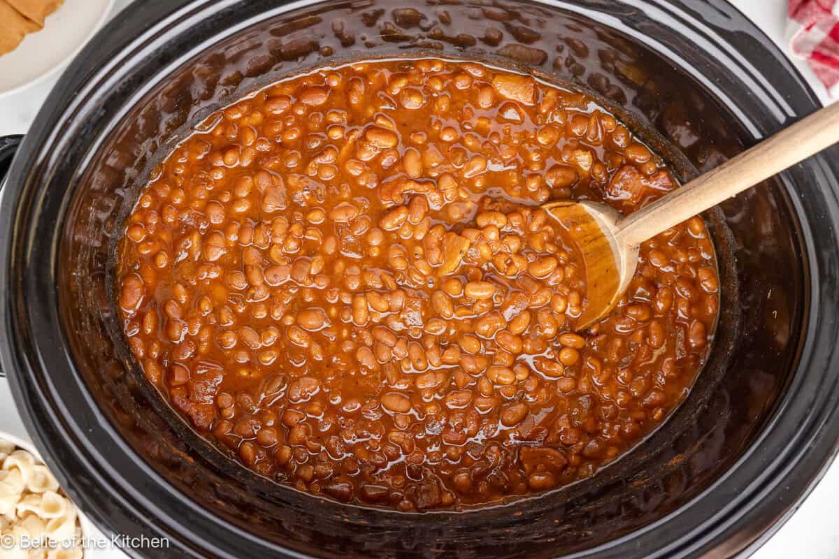 A black crockpot full of baked beans and a wooden spoon.