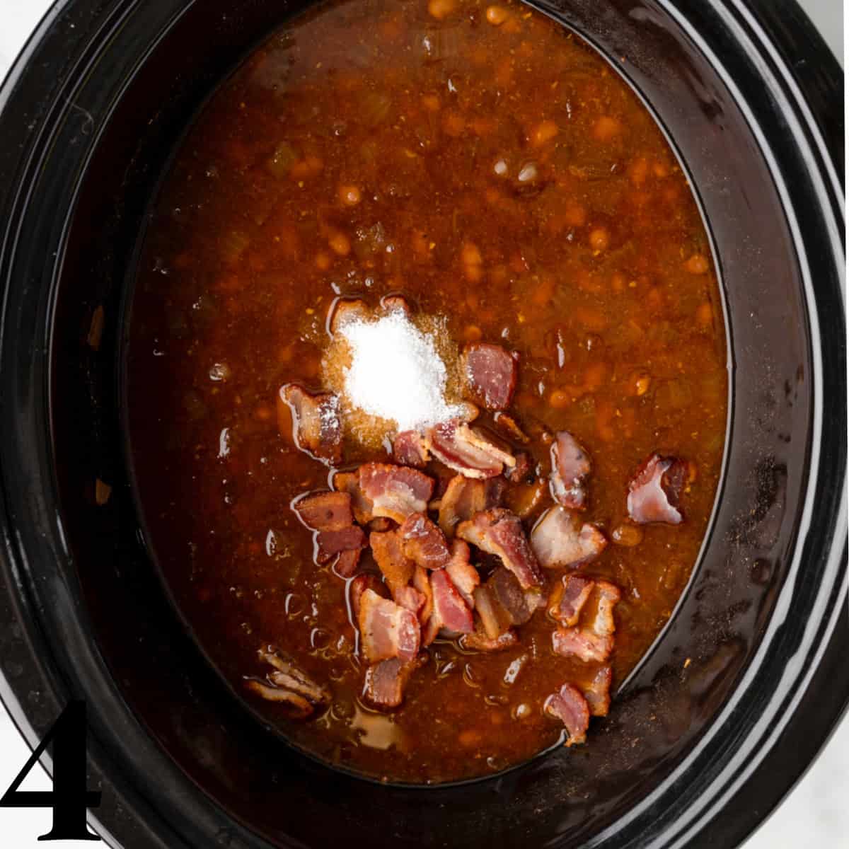 A black crockpot full of baked beans with salt and chopped bacon on top.