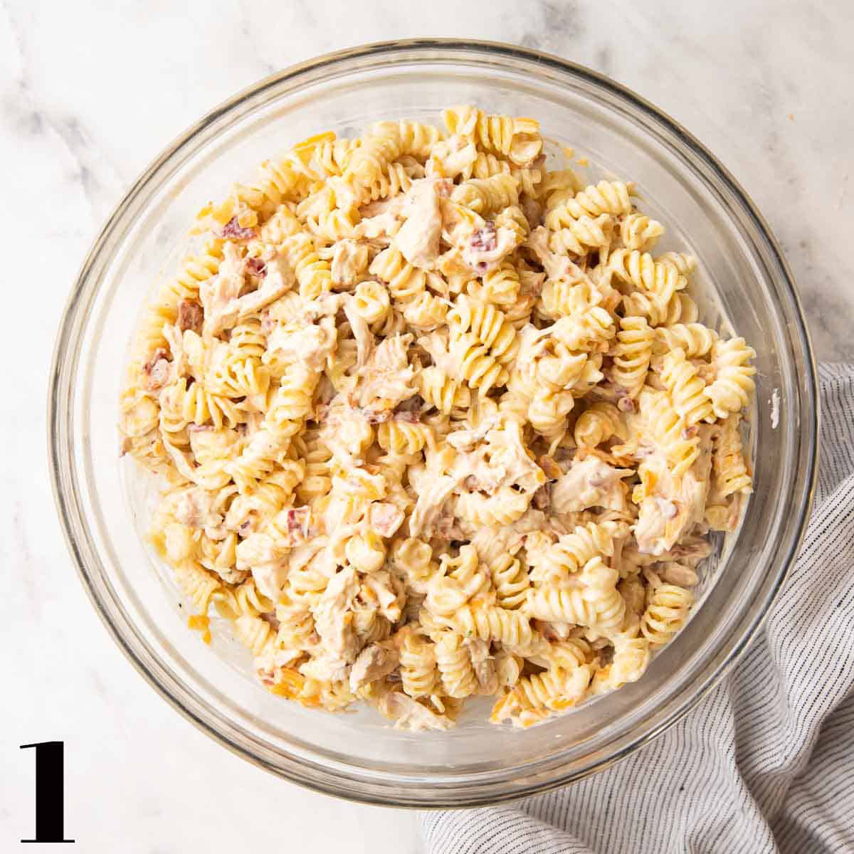 A glass bowl of pasta, chicken, sauce, bacon, and cheese.