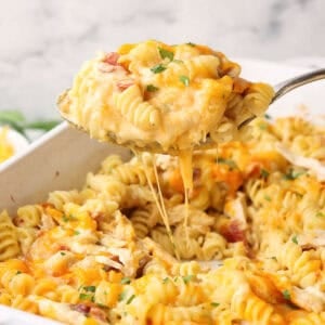 A spoon lifting food from a white baking dish full of pasta, chicken, bacon, and cheese.