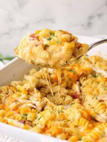 A spoon lifting food from a white baking dish full of pasta, chicken, bacon, and cheese.