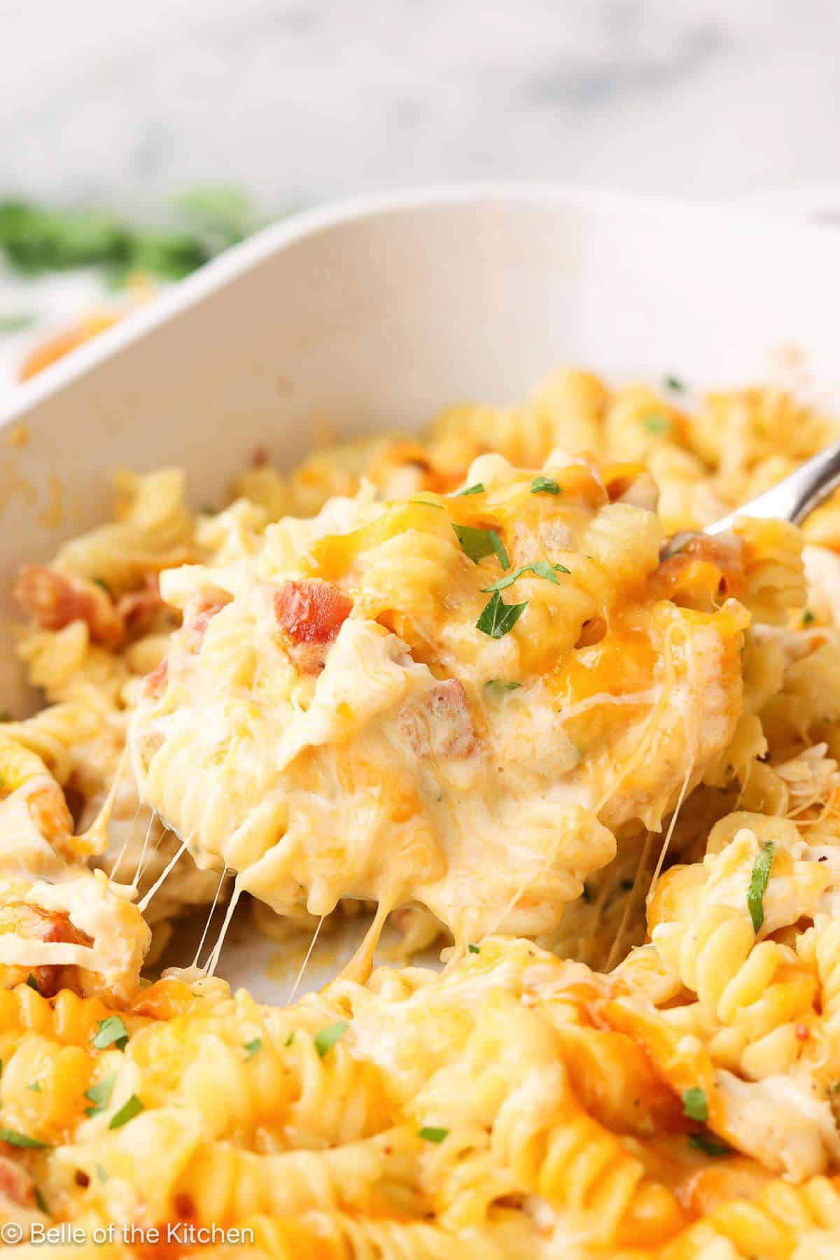 A spoon lifting food from a white baking dish full of pasta, chicken, bacon, and cheese.