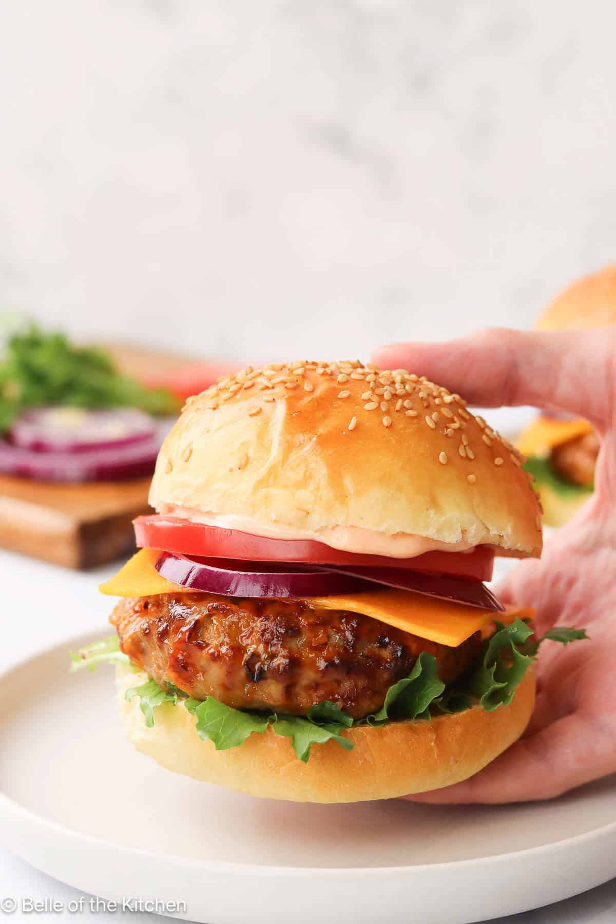 A hand holding a turkey burger with a bun and toppings above a white plate.