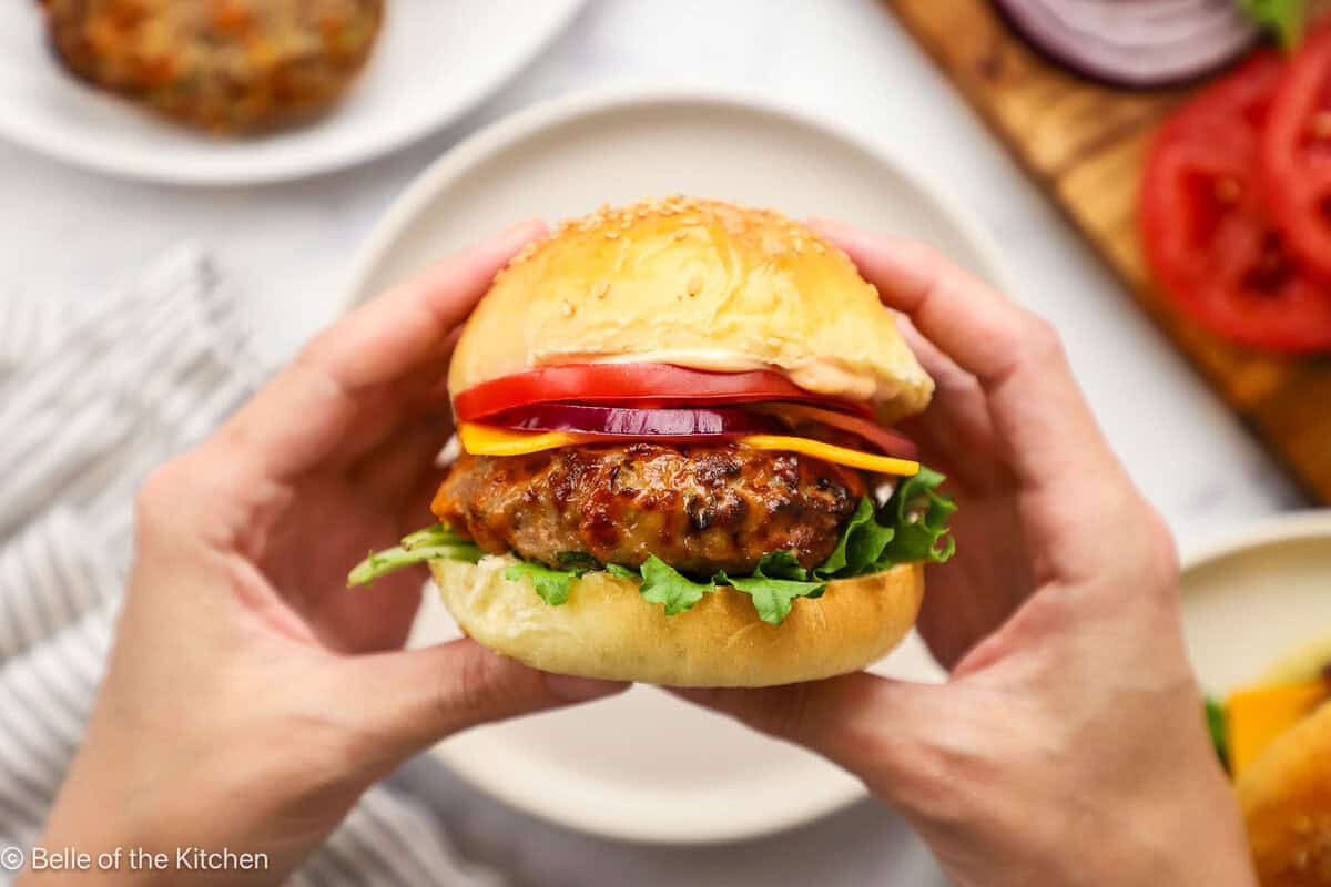 Hands holding a turkey burger with a bun and toppings above a white plate.