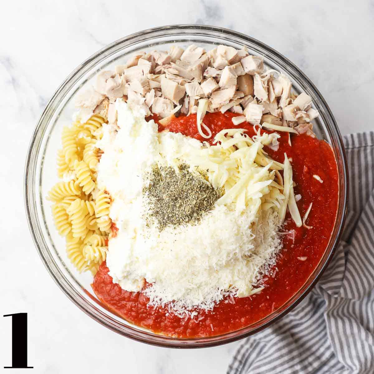 A glass bowl full of chicken, pasta, sauce, cheese, and spices.