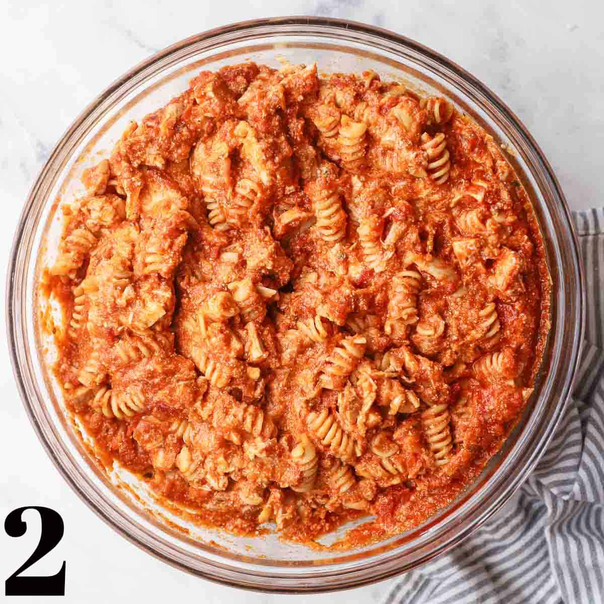 A glass bowl full of chicken, pasta, sauce, cheese, and spices mixed together.