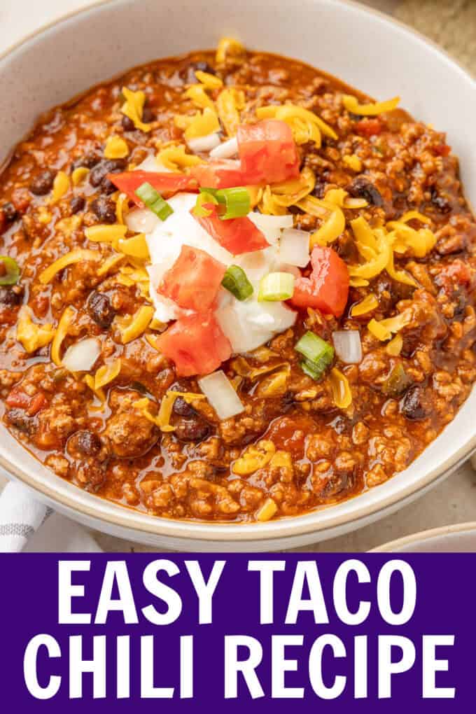 A white bowl of chili topped with sour cream, tomatoes, onions, and cheese.