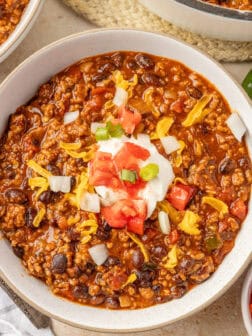 A white bowl of chili topped with sour cream, tomatoes, onions, and cheese.