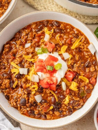 A white bowl of chili topped with sour cream, tomatoes, onions, and cheese.
