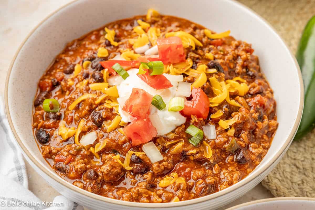 A white bowl of chili topped with sour cream, tomatoes, onions, and cheese.